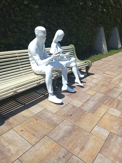 This image depicts two white, life-sized statues of a man and a woman seated on a light-colored park bench. The man is on the left, and the woman is on the right. Both statues are engaged in looking at or holding objects in their hands, suggesting they are reading or interacting with mobile devices. The bench has a green, ornate metal frame.

The statues are positioned on a paved walkway made of rectangular, multi-colored stone tiles in shades of brown, tan, and gray. To the left of the bench, the ground is partially covered by the bench's shadow, which forms distinct linear patterns. Behind the bench, there is a dense green hedge acting as a backdrop. Two triangular, light-colored concrete structures are visible behind the hedge, to the right of the statues. The environment suggests an outdoor public space, possibly a park or a promenade.

There is no information available to confirm or deny the location in the city of Oryol-Izumrud, Russia.