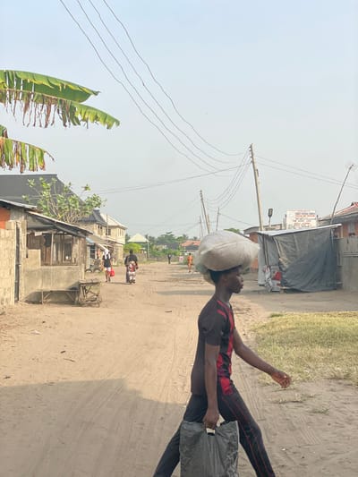 This image depicts a daytime scene on an unpaved, sandy road in Mosafejaw, Nigeria. The central figure is a young male, seen in profile, walking from right to left across the frame. He carries a large, light-colored sack or bundle balanced on his head and a dark, possibly plastic, shopping bag in his right hand. He wears a dark t-shirt with red accents and dark trousers with red piping.
In the mid-ground, other individuals are visible on the road: a person in a white top walking left with a red bag, and behind them, a person in a patterned dress walking away from the camera. Further down the road, a person on a motorcycle travels away from the viewer, and several distant figures are also present.
The environment includes single-story and multi-story buildings lining both sides of the road. On the left, a concrete block structure adjoins a wooden stall with a corrugated roof, featuring a wooden bench in front. On the right, utility poles with multiple overhead wires run along the roadside. A white sign for "CHRIST APOSTOLIC CHURCH" with "MOSHAFEJA BRANCH" is legible in the distance on the right, above a structure covered by a dark tarp. Overhanging banana leaves are visible in the upper left, and the sky is a hazy, light blue or grey. Long shadows across the road suggest morning or late afternoon light.