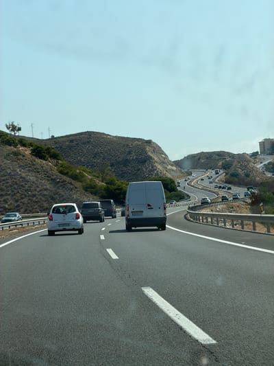 The image shows a view from a vehicle traveling on a multi-lane highway. The highway curves to the right, with rolling, dry hills on either side, dotted with sparse vegetation. Several vehicles are visible on the highway, including a white hatchback in the foreground, a dark SUV and a dark van further ahead, and a white van on the right side of the frame. The sky is clear and pale blue. In the distance, a multi-story building is visible on a hilltop to the right. The scene appears to be in a warm, dry climate.