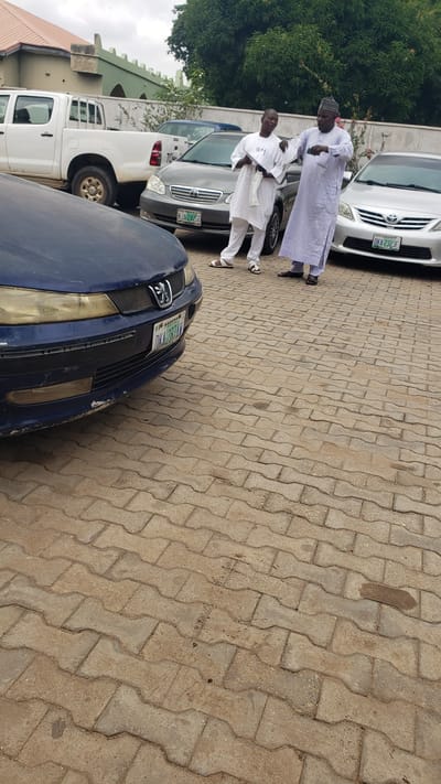 The image shows an outdoor scene in Unguwar Shanu, Nigeria, with two men standing next to parked cars on a paved surface. The man on the left is wearing a white kaftan and a white cap, holding a piece of paper. The man on the right is wearing a light purple kaftan and a dark cap, appearing to be taking a photo with his phone. In the background, there are several cars including a white pickup truck, a silver Toyota sedan with license plate "OKA-232JF", and a dark blue sedan with license plate "OKA 297AD" visible on the front. The environment appears to be a parking area with a building and trees in the background. The overall lighting suggests an overcast day.