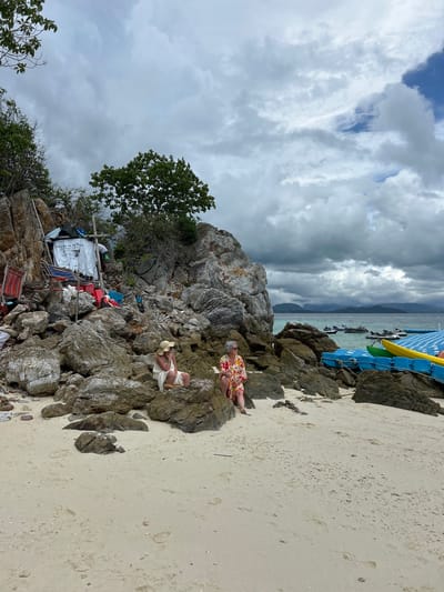 Two individuals are seated on rocks on a sandy beach in Phru Nai, Thailand. The person on the left wears a white dress and a straw hat, and the person on the right wears a floral print dress. Behind them, a rocky cliff face rises, adorned with makeshift structures and various items, including what appears to be a sign that reads "Welcome to My Paradise." The ocean is visible in the background, with several jet skis and floating platforms in the water. The sky is overcast with dramatic clouds. The scene suggests a somewhat rustic or off-the-beaten-path tourist destination.