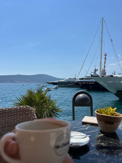 The image depicts a close-up of a person holding a coffee cup in the foreground, with a blurred background of a harbor. The table in front of the viewer has a black marble surface, a small potted succulent, a menu, and a saucer with a spoon and sugar packet. To the left, a woven chair and a palm frond are visible. The middle ground shows a body of blue water, possibly a bay, dotted with small boats. In the background, a line of large yachts and sailboats are docked. A mountain range forms the horizon. The sky is clear and blue. The scene is situated in Donja Lastva, Montenegro, a coastal town known for its marina.