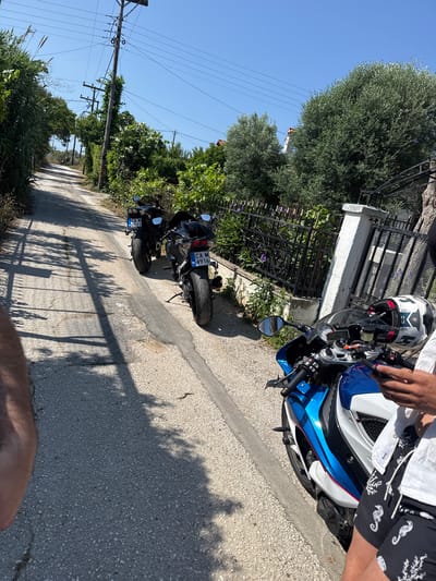 The image captures a sunny outdoor scene on a paved road, likely in a rural or semi-urban setting like Agia Triada, Greece. In the foreground, a person is standing next to a blue, white, and black sportbike, possibly a BMW S1000RR, with their right hand on the handlebar. The person wears a white short-sleeved shirt and black shorts adorned with white seahorse and coral patterns. A white helmet with black and red accents (possibly a Shark brand) rests on the motorcycle's tank.
Further down the road, two black motorcycles are parked on the right side. One of these bikes clearly displays a Bulgarian license plate: "CA M 4916 BG". The other black motorcycle has a partially visible plate, "CA M 0511 BG".
The road is bordered by lush green vegetation, including tall trees (potentially olive trees) and dense bushes. Wooden utility poles with power lines run along the left side of the road. On the right, a low white wall with a black metal fence separates the road from more greenery.
The time of day appears to be midday or early afternoon, as evidenced by the clear blue sky and the strong, distinct shadows of trees and the fence cast across the asphalt. The scene suggests a break during a motorcycle trip or an outing.