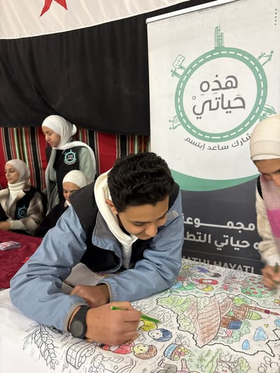 The scene depicts an indoor activity in Kafr Batna, Syria, involving multiple individuals. In the foreground, a young male, approximately early teens, with dark, wavy hair, is leaning over a large, uncolored line-art drawing spread across a white table surface. He wears a light blue zippered jacket over a white hooded sweatshirt and a black digital watch on his left wrist. His right hand holds a green marker, actively applying color to the intricate drawing, which illustrates a landscape featuring stylized buildings, trees, human figures, and baskets of produce. Partially visible to the male's right, a second individual, wearing a light-colored headscarf and a pink scarf, also uses a green marker to color the same drawing.
In the mid-ground, to the left, three young females are present. One stands, wearing a white hijab and a light green shirt under a black vest adorned with a circular logo containing Arabic script; she is looking towards the left. Another female is seated at a red-topped table, similarly dressed in a white hijab and a cream-colored top beneath a checkered jacket with the same logo; she looks generally forward. A third female, also wearing a white hijab, is partially visible behind the seated individual.
The background includes a large vertical banner on the right side, featuring a prominent green circular logo. The logo contains Arabic text centrally, reading "هذه حياتي" (This is my life), with "شارك ساعد ابتسم" (Share, help, smile) below it. The circle's perimeter is decorated with small icons representing buildings, people, and community elements. To the left, a fabric backdrop displays a white section with a red star at the top, followed by a solid black section, and a lower striped pattern of black, red, and beige.