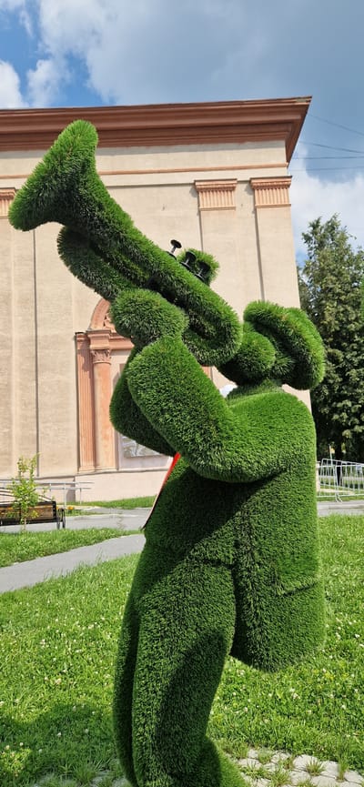The image shows a topiary sculpture of a trumpet player, made entirely of lush green grass. The sculpture is positioned outdoors in a grassy area, with a light-colored building with brown trim and architectural details in the background. A pathway and a bench are visible to the left of the sculpture, and trees and a white fence are in the distance. The sky above is partly cloudy and blue. The sculpture depicts a figure holding and playing a trumpet, with its body and limbs shaped from the dense greenery.