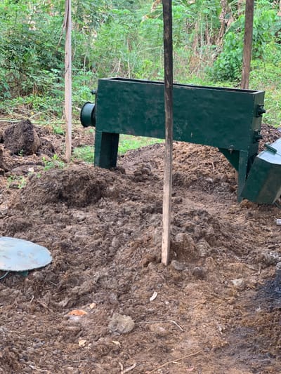 The image depicts a green metal structure, likely a composting or manure processing device, situated in a densely vegetated area. The structure rests on a bed of brown organic material, likely a mixture of manure and other compostable waste. A wooden pole is positioned diagonally across the frame, partially obscured by the vegetation. The structure is located in a rural area of Egbeda, Nigeria, at coordinates 7.119 latitude, 5.362 longitude.