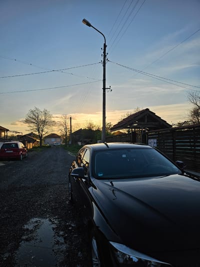 The image shows an outdoor scene in Krasnovo, Bulgaria. A black car is parked to the right. To the left of the car is a gravel road and a red car further down the road. A street light and utility pole are visible along the left side of the frame, stretching up towards the sky. In the background, there are trees, a house, and a wooden structure. The sky displays a gradient of colors, suggesting either sunset or sunrise. The road appears wet, indicating recent rain.