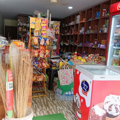 The image captures the interior of a small retail store in what can be inferred as Oxel, India. Dominating the foreground is a bunch of brooms in a bucket on the left, partially obscuring a metal display rack packed with various flavors of Lay's potato chips and Kurkure snacks. Behind the snacks, there is a black table filled with eggs, Ziggy snack box, and a shopping bag adorned with floral patterns.
To the right, a red and white freezer, bearing the brand name "Haier" and "Dairy Day" logo, is visible. The interior shelves of the store are densely stocked with a wide array of products, including snacks, household goods, and groceries. A Coca-Cola refrigerator can be seen on the right side.
The overall impression is that of a compact, well-stocked local store with a focus on snack foods and chilled items. The store exhibits the character of a typical small business found in urban India.