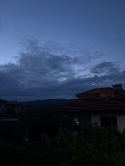 The image depicts a view of a building and a tree against a dusky sky filled with dramatic, dark clouds. The building appears to be a house with a tiled roof, and a solar water heater is visible on the roof. A dense, leafy tree dominates the right side of the frame, casting much of the foreground into shadow. In the background, a mountain range is dimly visible under the heavy cloud cover. A single, faint light can be seen on the mountain's slope. The overall mood is somber and somewhat mysterious, possibly indicating an approaching storm or the transition from day to night. The lighting suggests it is either dusk or dawn. The image seems to be taken in a residential area, given the presence of the building and the tree.