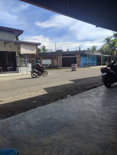 In the city of Cotglumpang, Indonesia, a person wearing a brown jacket and a dark helmet rides a motorcycle down a paved road. The motorcycle is dark gray with silver accents on the exhaust and wheels. The rider's jacket has a logo with the letters "MCI" and some illegible text in white and orange. The road surface is rough and appears to be asphalt with patches of gravel. To the left of the road, a building with a gray roof and white walls is visible. This building has a glass-fronted kiosk. Further down the road, there are more buildings with corrugated metal roofs and wooden structures. One building has blue doors and a sign with text that is too small to read. A red and white striped traffic barrel is placed on the side of the road in front of a building. A dark-colored motorcycle is parked on the right side of the image in the foreground. The sky is blue with scattered white clouds. Palm trees are visible in the background. The ground in the foreground is a concrete surface.
