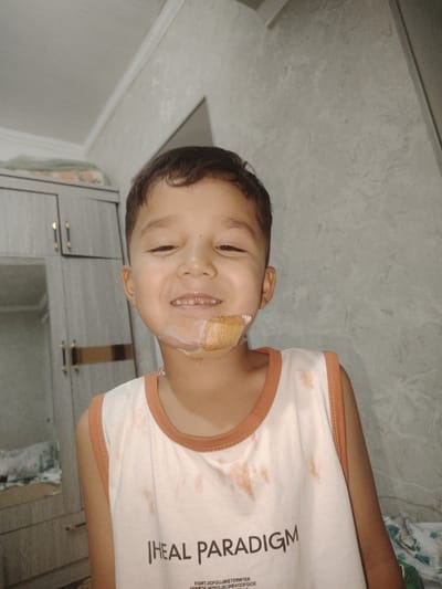 The image is a medium shot of a young boy in a brightly lit room. The boy is smiling and looking at the camera. He has a small bandage on his chin and some white substance around his mouth. His hair is dark and short, and he is wearing a white tank top with an orange trim and an orange collar. The tank top has some light brown stains on it. There is text printed on the tank top. In the background, to the left of the boy, is a large, light-colored wardrobe with two doors. The walls of the room appear to be textured and light gray in color. The ceiling is visible at the top of the image, with white molding. The overall impression is of an indoor setting, possibly a home. The location of Xonobod City, Uzbekistan is not visually identifiable in the image itself.