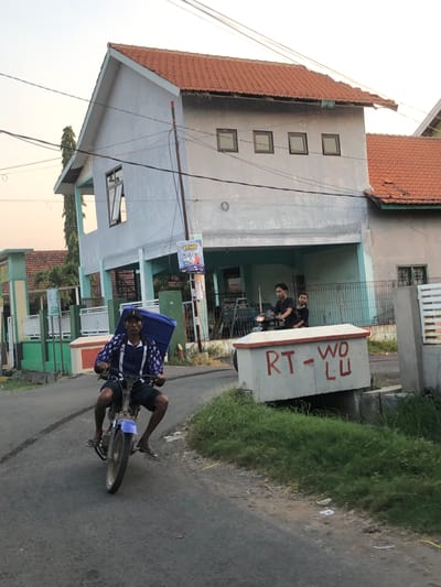 In Sentul, Indonesia, at an outdoor setting, a man is riding a motorcycle on a paved road. He is wearing a dark blue jacket with a white star pattern, dark shorts, and flip-flops. On the back of the motorcycle is a large blue box. Behind the man, two young men are riding on another motorcycle. They are both wearing dark shirts. In the background, there is a two-story house with a red tiled roof and light blue walls. A banner with pictures of people is hanging on a pole in front of the house. A concrete structure with red lettering that reads "RT - WO LU" is situated to the right of the road. The environment is a residential area with green grass and some trees visible. The sky is a pale orange, indicating either early morning or late afternoon.