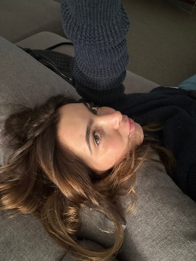 A single light-skinned adult female is depicted from an overhead perspective, positioned in an indoor setting. She has brown hair and light-colored eyes, and her head is resting on a grey, upholstered surface, likely a sofa or bed. She is wearing a dark, possibly navy or charcoal, textured knit garment, consistent with a sweater or jacket, which features a metallic zipper closure. Her right arm (from the viewer's perspective) is raised, indicating she is holding a device, potentially a camera or smartphone, to capture the image. A black, woven-texture bag or clutch with a metallic zipper is visible on the grey upholstered surface to her left. The background is dark and out of focus, suggesting subdued lighting. The specific location within Nogueira e Silva Escura, Portugal, cannot be identified from the visual content of the image.