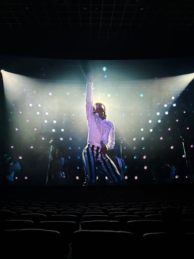 A person, likely a performer, stands on a stage illuminated by a bright spotlight. The person is wearing a sparkly white top and striped pants. The stage is decorated with numerous small, colored lights, creating a backdrop. The right side of the image is dark, possibly showing audience seating or stage equipment. The location is likely in Taihe District, China.