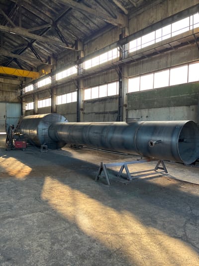 An industrial interior scene displays a large, cylindrical metal tank, likely for storage or processing, positioned horizontally on metal stands. The tank is the primary subject, occupying the center of the frame and extending towards the right. It is a dark, metallic color with visible seams and fittings. To the left of the tank, a welding machine and equipment are present, suggesting ongoing or recent fabrication work. The environment appears to be a workshop or factory floor, characterized by concrete ground, exposed ceiling beams, and large windows that allow sunlight to stream in, casting long shadows. The overall impression is one of heavy industry and manufacturing.