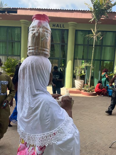 The image depicts an outdoor scene in Madina, Ghana, featuring a gathering near a building identified as a "HALL." In the foreground, a woman is seen from behind, balancing a large clear plastic jar with a red lid on her head. The jar contains numerous individually wrapped brown cylindrical food items. She wears a white head covering embellished with rhinestones, a white garment with a white lace-trimmed collar, and a floral-patterned skirt. Her hands hold a light-colored object, possibly a small bag or folded paper.
The background features a light green building with prominent glass windows and several light-colored pillars. The word "HALL" is visible in dark letters on its facade, beneath a reddish-brown roof. The ground is paved with reddish-brown interlocking bricks. To the right of the main subject, a man in a dark shirt with "CARRY" visible is looking down at a mobile phone. Further right, two women are seated on the paved ground near a planted area; one wears red, the other blue. Another person in a green dress stands behind them. Sparse vegetation, including potted plants and a slender palm tree, are visible around the building. The scene is brightly lit by natural daylight. Partially visible on the far left is a person wearing an olive green patterned tunic.
