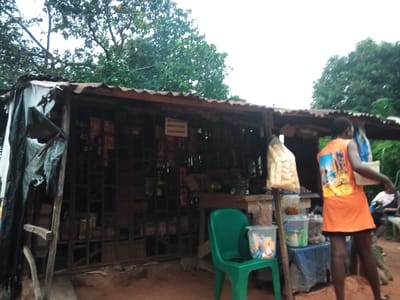 A medium shot depicts an outdoor market stall under a corrugated iron roof with a sagging front awning covered in plastic sheeting. The stall is filled with various goods. A sign with text in what appears to be French is visible. To the right of the stall, a young Black female wearing an orange t-shirt with a graphic design on the back and a black skirt stands, holding a white bag and a white container with a blue lid. A green plastic chair is next to her, holding a clear plastic container filled with small items. In the background, more stalls and foliage are visible, suggesting a rural or semi-urban setting. The ground is dirt. This scene is located in Nanka, Nigeria.