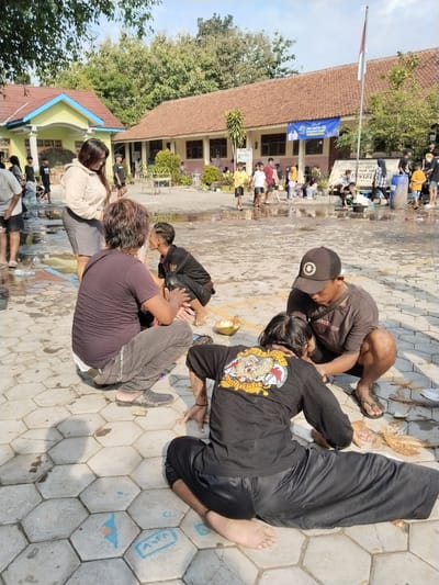 The image shows a group of people in an outdoor setting, likely in Karangmalang, Indonesia, given the banner in the background mentioning the area. In the foreground, several individuals are crouched on a paved surface, possibly involved in some activity. One person is wearing a black shirt with a graphic design on the back and black pants, sitting with their legs spread out. Another individual is wearing a brown t-shirt and a cap, appearing to be focused on the person in black. Another individual with dark hair is crouched nearby, and a woman in a light-colored hoodie and grey shorts is standing in the background. There are halved coconuts on the ground near the group, and there seems to be water on the paved surface. In the background, there are more people, possibly children, walking or playing in a wet area. Buildings are visible in the distance, including one with a tiled roof and another painted yellow with blue trim. A flag is flying in the background.