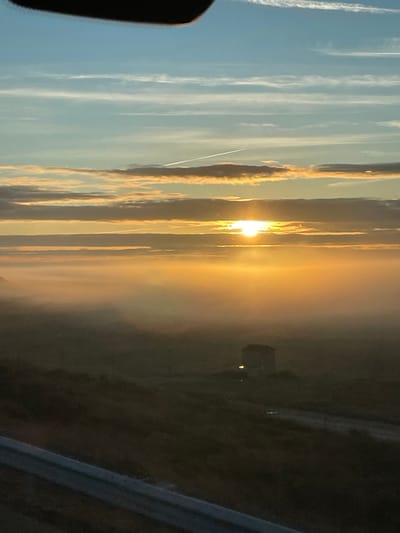 The image captures a vibrant sunset. Streaky clouds dominate the left side of the frame, with an airplane contrail cutting across. The sun bursts through the clouds, casting a golden glow. Hazy, golden light obscures a building in the distance. A dark foreground and a car window and rail are visible. The image appears to be taken from inside a car.