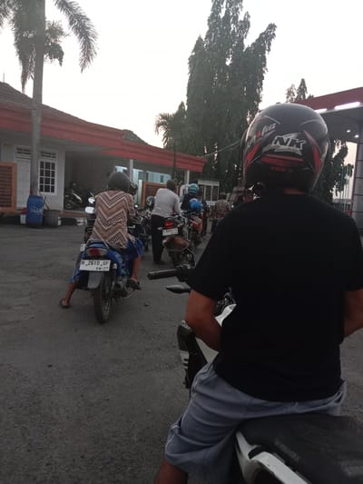 In Brangsong, Indonesia, at dusk, several individuals are lined up on motorcycles on a dirt road in front of a building with a red awning. A person wearing a black shirt and grey shorts, with a black and red helmet, is facing away from the camera, seated on a motorcycle in the foreground. Behind them, another person in a patterned shirt and blue pants is on a blue motorcycle, facing left. Further back, people on other motorcycles are visible. Tall trees are in the background, and a few palm trees are to the left of the building. The sky is bright white, suggesting overcast conditions or early dawn/late dusk. The overall scene depicts a casual gathering of motorcyclists, possibly waiting for something or preparing to depart.