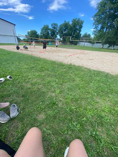 This is a vertical, eye-level shot of a group of people playing volleyball on a sand court on a sunny day. The sky is a clear, bright blue with a few scattered white clouds. Lush green trees line the edge of the court, and a white building is visible in the lower left corner. The sand court has red boundary lines.

Several individuals are engaged in the game, with some actively playing and others watching. One person in a black shirt and shorts is mid-action on the sand court, while another in a white shirt and shorts is also playing. There are other figures in the background, some near the net.

In the foreground, the perspective shifts to a grassy area. A person's legs and feet are visible, with a pair of grey sneakers lying on the grass. Another pair of white shoes is also on the grass nearby. The time of day appears to be daytime, judging by the bright sunlight and clear sky. The overall atmosphere seems to be one of leisure and outdoor recreation. No text is visible in the image. The location context is Mostovskoy, Russia.