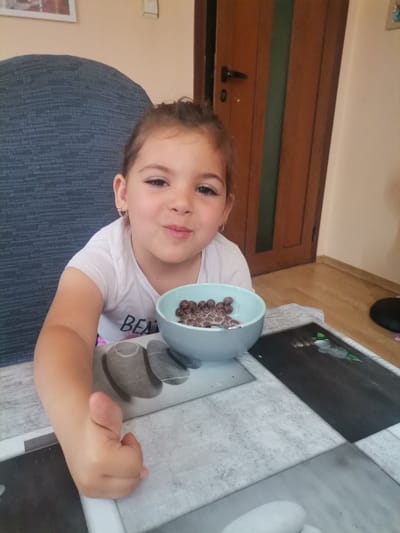 A young female child, with brown hair pulled back, is seated at a table, looking directly at the camera and exhibiting a faint smile, in Obrochishte, Bulgaria. She is wearing a white short-sleeved t-shirt with partially visible lettering ("BEA") and small gold-colored hoop earrings with pink heart accents. Her right arm is extended towards the camera, with the thumb raised in a positive gesture. A light blue bowl containing dark brown spherical cereal and milk, with a spoon immersed, rests on the table in front of her. The table surface features a patterned placemat or tablecloth with grey and white stone-like or tiled sections, and a dark rectangular panel with a white floral and green leaf motif. Behind the child, a dark blue textured chair back is visible. The background consists of a light beige wall, a brown wooden door with a dark handle, and a light brown wooden floor.