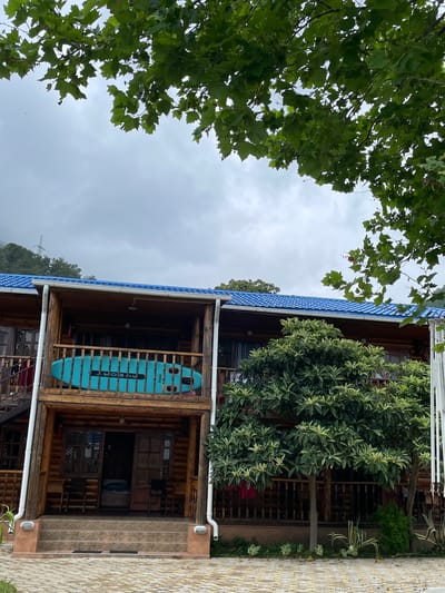 In Abaata, Abkhazia, a wooden log cabin with a blue tiled roof stands under a cloudy sky. A teal-colored paddleboard with black lettering is propped on the railing of the second-story balcony. The cabin has multiple levels with wooden balconies and staircases. Green trees and foliage surround the building, and a cobblestone path leads to the entrance. The scene appears to be a resort or a vacation rental.