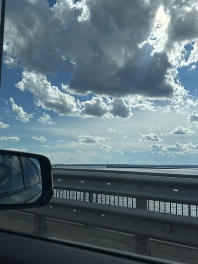 This media file captures a view from a vehicle on a sunny day, likely during daytime, with a clear blue sky dotted with fluffy white and dark gray clouds. The scene appears to be a bridge or overpass, indicated by the presence of a metal guardrail on the right side of the frame, showing a repeating pattern of horizontal slats. The guardrail is made of thick metal poles and a textured surface. Beyond the guardrail, a body of water is visible, possibly a river or lake, reflecting the sky. In the distance, a faint outline of buildings suggests an urban or suburban area.
The foreground of the image features a car's side mirror, reflecting the sky and clouds, as well as the interior of the vehicle. The reflection in the mirror is slightly distorted. The overall impression is a scenic drive, with the vast expanse of the sky and the contrasting metallic structure of the bridge. There is no visible text in the image. The weather appears fair, with sunlight breaking through some of the clouds, creating dramatic shadows and highlights. The location context provided is Пальники, Russia.