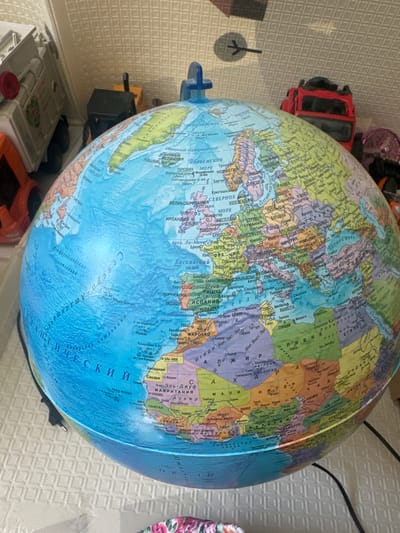 A close-up, slightly angled shot shows a large, terrestrial globe positioned on a patterned white surface. The globe displays continents and oceans with visible country borders and place names, predominantly in Cyrillic script. Parts of toys are visible in the background, including a white truck with a red accent on the left and a red truck on the right. The environment appears to be indoors, with a textured wall in the background. The lighting is diffuse, casting soft shadows. There is no discernible connection to the city of Novoselye, Russia, in the visual information provided.