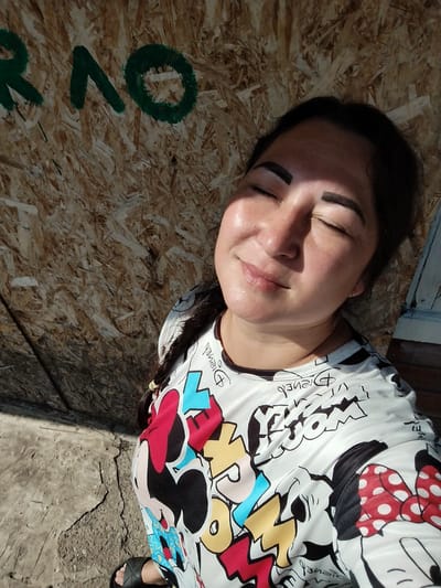 A woman is captured in a close-up, high-angle selfie outdoors in Krasny Gulyai, Russia. She is standing in front of a wall made of oriented strand board (OSB) with green painted letters that appear to spell "RAO." The wall is rough and textured, showing the wood chips embedded within it. The ground beneath her is concrete, with visible cracks. Sunlight is casting a strong shadow from her left side, highlighting the contours of her face. She has her eyes closed, her head tilted back slightly, and a subtle smile on her lips, suggesting she is enjoying the sun. Her dark hair is pulled back, and a braid is visible over her shoulder. She is wearing a white long-sleeved shirt adorned with a graphic print featuring characters and text, possibly from Disney, including Minnie Mouse. Her left foot, wearing a black sandal, is visible at the bottom of the frame. The scene appears to be in a somewhat unkempt outdoor area, possibly a construction site or an abandoned space.