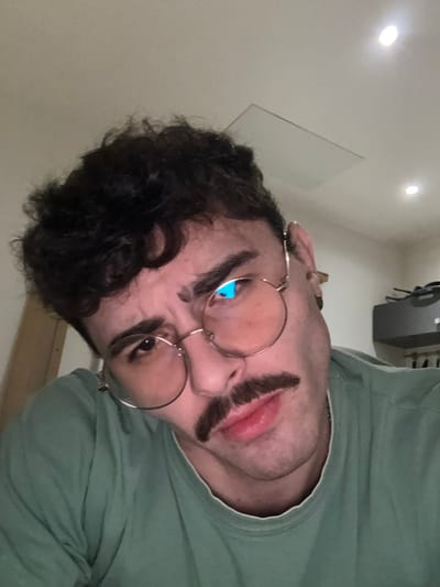 A person with dark curly hair, round glasses, and a thick, fake mustache is angled in the frame. The person wears a light green shirt. Above the person, three ceiling lights are visible, along with a white board and a gray cabinet. The picture is taken from a low angle, making the person's face the focal point.