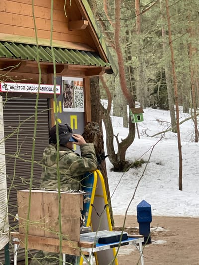 The image depicts an outdoor scene in Morskoe, Russia, characterized by a partial snow cover and a wooded environment. To the left, a light brown wooden building with a dark green corrugated roof displays two signs: one stating "ВЕДЕТСЯ ВИДЕО НАБЛЮДЕНИЕ" (Video Surveillance in progress) and another featuring "НАШ КАНАЛ" (Our Channel) with an owl image and two QR codes. A dark grey roll-down security shutter is partially visible below the signs.

In the midground, an individual is observed from the rear-left, wearing a camouflage jacket and a dark cap. This person is holding a dark-colored smartphone to their face, appearing to capture an image or video. A large, brown, mottled owl is perched on the individual's gloved left forearm, facing slightly away from the individual. A yellow metal A-frame ladder or stand is positioned near the individual and the owl.

In the lower foreground, a portable table holds a wooden box-like structure and a clear plastic organizer. To the right, a blue conical post stands partially visible. The background comprises a sparse forest of pine and bare deciduous trees, with a small green house-shaped birdhouse attached to a tree trunk towards the center-right. Patches of snow cover the ground throughout the scene, indicative of cold weather conditions.