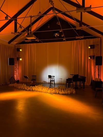 The image depicts an indoor performance space, likely a hall or theater, bathed in warm, orange lighting. The ceiling features exposed beams and industrial lighting fixtures, including a projector suspended from the center. The walls are draped with large, light-colored curtains, creating a soft backdrop.
In the foreground, the floor is covered with a multitude of unscented, battery-operated pillar candles, arranged in a dense, curving pattern that stretches across the space. The candles are illuminated by the ambient lighting and appear to be unlit.
Further into the scene, several objects are set up for a performance. To the left, a tall speaker stands on a tripod, positioned next to a lighting truss with two yellow spotlights. A music stand is also visible in this area. In the center, a cello rests against a chair, with another music stand beside it. Dominating the right-center of the image is a black grand piano. Behind the piano, another lighting truss with spotlights and a speaker is present, and a stack of candles is placed on a dark box. Another chair and a music stand are visible further back on the right. The overall impression is one of a stage set for a musical or atmospheric performance, with a focus on visual ambiance created by the lighting and candles. There are no people visible in the image. The location is not specified in the image or provided context.
