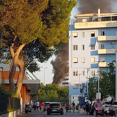 The image captures an urban street scene in Pescara, Italy, under daylight conditions, dominated by evidence of a structural fire. In the foreground on the left, a large, mature tree with a robust, textured trunk and dense green foliage partially obscures the view. Behind this tree, a portion of an orange-colored building with a white roof and a visible white cross emblem on its facade is present.

The midground features a paved street or road. A dark-colored car is positioned on this road, facing away from the viewer. Numerous pedestrians are visible on both the street and the sidewalk, walking in various directions; some appear to be in small groups, while one individual on the far right is depicted pushing a shopping cart.

In the background, a prominent multi-story building, light blue in color, stands on the right side of the frame. This building features multiple windows, some with exterior shades. A dense, dark gray to black column of smoke is actively billowing upwards from behind or within this light blue structure, rising well above its roofline, indicating an active fire. Additional green trees and foliage are visible in the background between the buildings. Multiple streetlights on tall poles are also scattered throughout the scene.