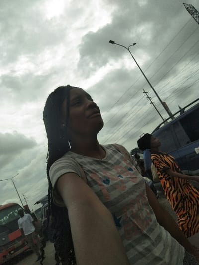 The image captures an outdoor urban scene in Ajah, Nigeria, under a heavily overcast sky. In the foreground, a young Black female with long dark braided hair and a grey and white striped t-shirt featuring pink hearts and blue text is depicted. Her right arm is extended, suggesting she is taking a selfie, and she is looking upwards and slightly to her right.
In the midground, to the right of the primary subject, another young Black female is visible, wearing a black head covering and an orange and black zebra-print dress, also looking upwards. Behind her is a dark blue SUV or van. To the far left, a red and white bus or truck is partially visible, with a man in a white t-shirt standing nearby. Overhead, multiple utility poles with numerous power lines crisscross the frame horizontally, and two streetlights are discernible, one prominently curved in the upper right. A tall metal lattice tower is also visible in the far upper right background.