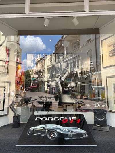This is a photograph of a shop window displaying artwork and merchandise. The window reflects the street scene outside, which includes buildings, trees, and a cloudy sky, suggesting it's daytime.
The primary subjects in the window display are two pieces of art. One is a large, glossy print of a silver Porsche 911, accompanied by the text "PORSCHE 911" in red and white. To its left, there's a black and white photographic print of a man in a suit leaning against a vintage motorcycle, with text reading "Dior" visible on a black background element.
Reflected in the window are street views of what appears to be a European city, characterized by multi-story buildings with arched windows and balconies. The street is paved with cobblestones and lined with trees. There are glimpses of people on the street, and outdoor seating areas, indicating a lively urban environment. The overall impression is one of a sophisticated retail space offering luxury items and art, likely in a historic European setting given the architectural reflections and the location context of Baden-Baden, Germany. The lighting and reflections suggest a bright, possibly sunny day, despite the visible clouds.