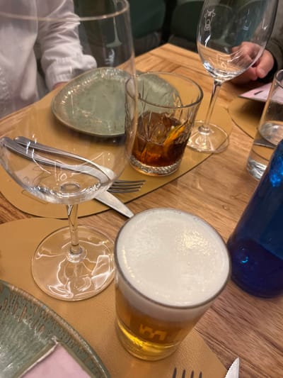 The scene is a close-up view of a table set for four people at a restaurant in Las Palmas de Gran Canaria, Spain, located at the coordinates 28.102 latitude, -15.414 longitude. A beer glass with a white head is in the center of the image. Next to it is a blue glass bottle. To the left, there are two empty wine glasses and a partially visible plate with green patterns on it. The table is made of wood. The place setting is in a casual, yet relaxed dining environment.
