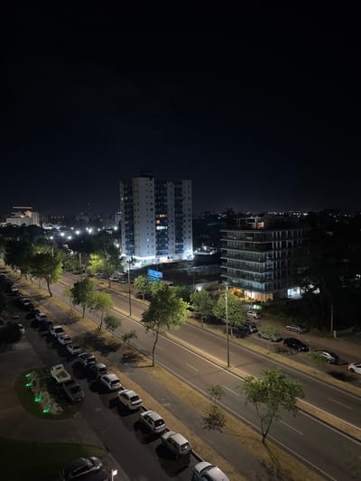 A nighttime aerial view shows two high-rise buildings, one with a modern, geometric design. A road runs parallel, lined with parked cars and trees illuminated by streetlights. The cityscape extends into the distance with a row of lights following the path of the road. The rest of the frame is dominated by darkness, highlighting the illuminated urban landscape.