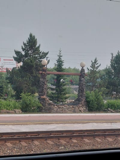 The image shows a stone archway with two large white globes on top, which appear to be lamps, with a wooden beam connecting them. The archway is situated in front of a line of trees, with a large billboard visible behind the trees to the left. The billboard features a portrait of a man and Russian text. In the foreground, there are train tracks, a platform, and some vegetation. The scene is outdoors and appears to be during the daytime under an overcast sky. This landmark is located in the city of Kargat, Russia.