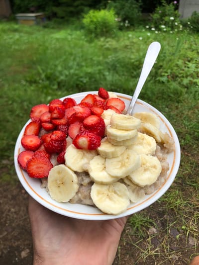 A white bowl of oatmeal topped with sliced strawberries and bananas is held in a person's hand. The bowl has an orange trim. A silver spoon rests in the oatmeal. The background is out of focus and shows green grass and foliage, with a hive visible in the distance. The image is taken outdoors, likely in a rural or garden setting. The approximate location is Svirstroy, Russia.