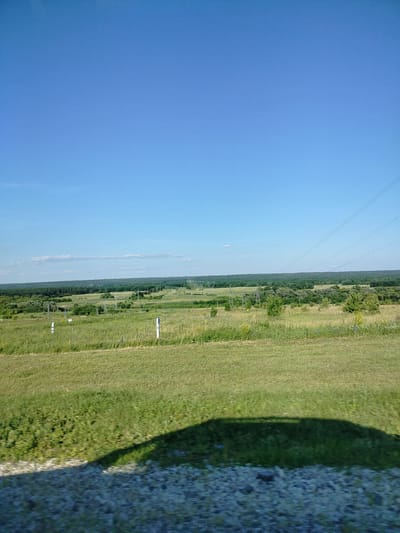 This media file captures a vast, open landscape under a clear blue sky, suggesting a daytime setting, likely mid-morning to late afternoon, with fair weather. The scene is dominated by rolling green fields, with a line of trees forming a dense forest in the middle ground and stretching towards the horizon. Interspersed within the fields and beyond are numerous electrical transmission towers and power lines, indicating infrastructure present in the area. The foreground shows a strip of dry, gravelly ground and a large, dark shadow cast by an unseen object, possibly a vehicle, suggesting the image was taken from a moving perspective.
There are no people visible in the image. The subjects are the natural landscape, the man-made power infrastructure, and the shadow in the foreground. The overall activity is observational, documenting a scenic yet utilitarian view of the countryside. Notable details include the distinct demarcation between the grassy fields and the dense forest, the repetitive structure of the power line supports, and the clear, unblemished sky. The presence of the shadow and the slight blur in the foreground suggests movement, possibly travel through the area. No text is visible in the media file. The context provided places this scene in Grabovo, Russia, contributing to the understanding of the rural, possibly Eastern European, setting.