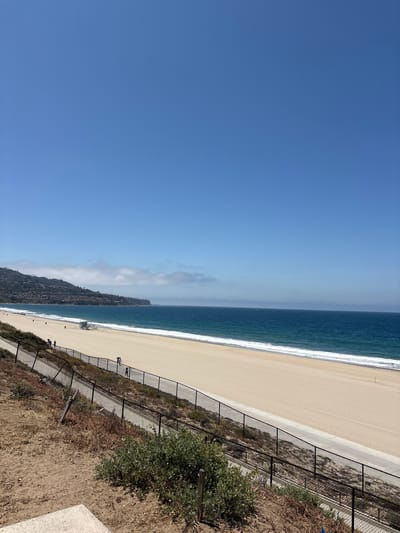 The image shows a view of a beach in Redondo Beach, California, USA. The sky is predominantly clear blue. The ocean is a deep blue color with white waves breaking along the shore. A wide sandy beach stretches along the coastline. A black chain-link fence runs parallel to the beach, separating it from a walkway. Beyond the walkway, there is a slope covered with dry vegetation and some greenery. Some people are walking along the beach. A hillside covered with vegetation and some buildings is visible in the distance, partially obscured by fog.
