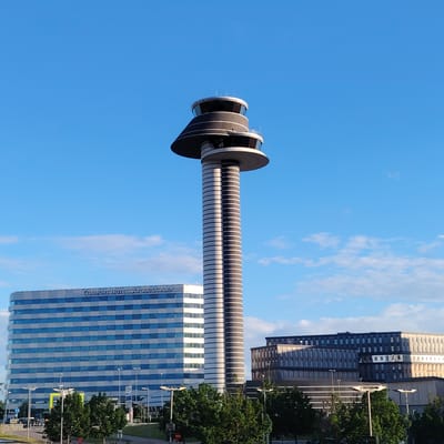 The image displays an architectural scene at Stockholm-Arlanda Airport in Sweden. The prominent central feature is a tall, slender air traffic control tower. Its cylindrical shaft is horizontally striated with alternating dark grey and light grey bands. The tower culminates in a multi-tiered top section featuring panoramic dark-tinted windows, a wide, flat, dark disc-like platform, and a smaller, lower platform with windows and a safety railing.
To the left of the tower, a large, multi-story building is visible, identified by "COMFORT HOTEL ARLANDA AIRPORT" text on its upper facade. This building has a rectangular form with a predominantly grey exterior interspersed with horizontal bands of reflective dark blue windows.
To the right of the tower, several other modern buildings with dark facades and multiple windows are present, exhibiting varying heights and rectilinear designs. Some surfaces of these buildings reflect sunlight.
The sky is a clear, bright blue with scattered, wispy white clouds, particularly concentrated in the lower portions of the sky. In the foreground, numerous green deciduous trees obscure the immediate ground level. Elements of infrastructure such as streetlights with multiple arms and a yellow structure (possibly signage or part of a parking facility) are partially visible among the trees and behind them. A road or paved area is discernible in the lower-left corner. The scene is uniformly illuminated by natural daylight, casting shadows that delineate the structures. No individuals are depicted in the image.
