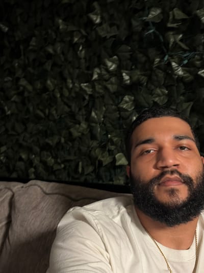 A man with dark hair and a full, well-groomed beard poses in front of a wall covered in artificial green leaves. He wears a light-colored long-sleeved shirt and a thin gold chain. His expression is neutral as he looks at the camera. The lighting is somewhat dim, casting shadows on his face and the background. A small portion of what appears to be a bed or blanket is visible in the bottom right corner.