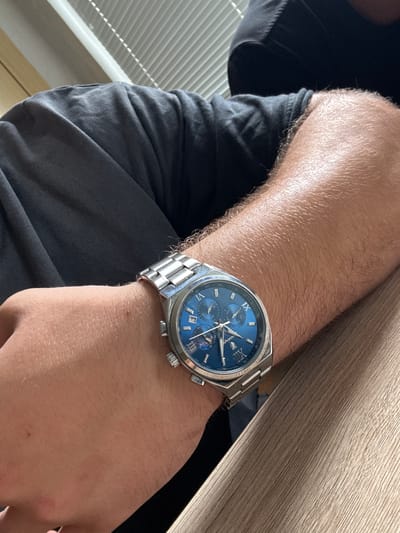 The image presents a close-up, high-angle view of a person's arm and hand, centered on a wristwatch, in an indoor setting located in Nova Zagora, Bulgaria. The arm, moderately covered with light brown hair, is positioned with the wrist resting on a light brown wooden surface, likely a desk or table. The person is wearing a dark grey or black t-shirt or casual garment, visible on the left side of the frame.
The primary object of focus is a silver-toned wristwatch featuring a metal link bracelet and a round case with three prominent pushers on its right side. The watch boasts a vibrant blue dial adorned with Roman numerals at the XII, IX, VI, and III o'clock positions, complemented by stick indices for the remaining hours. The dial integrates three subdials: one clearly indicating the day of the week, displaying "TUE" (Tuesday); another suggesting a 24-hour format with visible numbers such as 12, 18, and 24; and a third subdial with a less discernible function but showing numerical markings. The brand name "Poedagar" is distinctly visible below the 12 o'clock position, and "JAPAN MOVT" can be faintly read near the bottom of the dial. The watch's bezel includes a tachymeter scale with numbers such as 60, 65, 70, 80, 100, 120, 160, 185, and 200. The time displayed on the main hands appears to be approximately 10:25 to 10:28.
In the background, on the far left, white horizontal window blinds or grilles are partially visible, suggesting proximity to a window. The lighting is bright and indirect, indicative of daytime. The image solely captures the arm and watch, without revealing any specific activity beyond wearing the watch or conveying particular emotions.