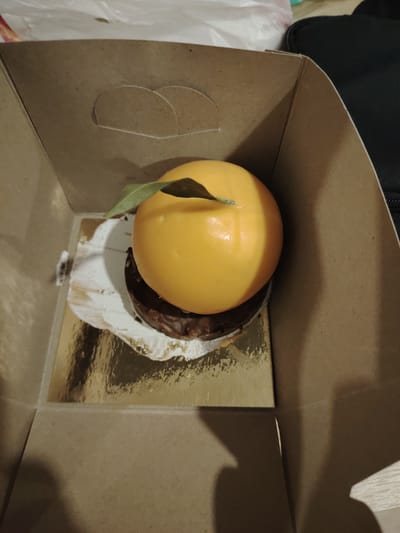 A vibrant, spherical dessert, resembling a large orange or mandarin, is the central subject of the image. It features a smooth, glossy orange exterior and is topped with a single, small green leaf, suggesting a fruit-inspired flavor, possibly citrus. This delicate confection rests upon a dark, round, ridged chocolate base, which appears to be a biscuit, cookie, or brownie.
The entire dessert assembly is placed on a shiny, reflective gold-colored cardboard disc, likely a disposable cake board. There are visible smudges of white cream or frosting on this gold disc, indicating some movement or handling of the dessert.
The dessert and its base are contained within an open, light brown cardboard box, typical for pastries or cakes. The box has folded walls and cutouts for a handle on the unseen top flap. The interior of the box provides a simple, neutral background, drawing focus to the colorful dessert.
In the background, outside the confines of the box, several blurred objects are visible. To the upper left, a crinkled white plastic material, possibly a bag, can be seen. To the upper right, a dark, likely black, fabric object, such as a bag or case, is partially in view. The bottom right corner shows a light-colored surface, possibly a table or countertop.
The lighting appears to be artificial and internal, creating subtle shadows within the box. No individuals are present, and there is no visible text. The scene captures a beautifully crafted dessert, implying it is either newly packaged or about to be enjoyed in an indoor setting in Tepebaşı, Turkey.