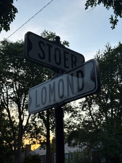 This is a low-angle, eye-level, medium shot of two street signs mounted on a pole, reading "STOER" and "LOMOND". The signs are black with silver lettering and are set against a backdrop of trees and a pale blue sky. The sun is setting, casting a warm orange glow through the trees. The overall lighting is dim, suggesting dusk. The scene appears to be located at an intersection of Stoer and Lomond roads in Shaker Heights, Ohio, United States of America.