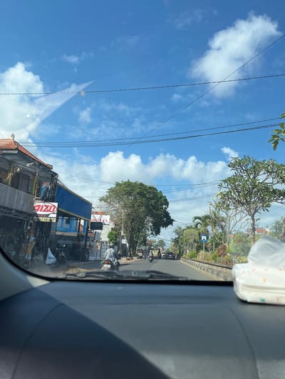 The image, taken from the interior of a vehicle, depicts a street scene in Jembrana, Indonesia, on a clear day with scattered clouds. The road is asphalt, with a concrete divider and embankment. The left side of the road features buildings with orange tiled roofs and signs, including one for "QIZO MAS HELMET" and another for "HOTEL CIPTA KARYA". A person on a motorbike is visible near the buildings. Further down the road, other vehicles, including cars and motorbikes, are present. Lush green trees and palm trees line the right side of the road. In the foreground, a box of facial tissues rests on the dashboard of the vehicle. Overhead, power lines crisscross the sky. The overall impression is a view of a street in a tropical or subtropical urban environment.