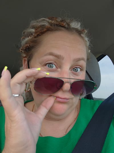 A woman is captured in a close-up, selfie-style shot, seemingly from inside a vehicle, with her head and shoulders filling the frame. Her curly, blonde and reddish-brown hair is styled in braids that frame her face. She is wearing a bright green shirt and a seatbelt across her chest. Her left hand is raised, with long, neon yellow nails, and she is holding a pair of aviator sunglasses, tilting them slightly downward with her thumb and index finger, revealing her blue-grey eyes. She is looking directly at the camera with a playful or curious expression, her lips slightly parted. A delicate gold chain necklace is visible around her neck. The background is blurred, suggesting movement, with a hint of a headrest and what appears to be a window. The lighting is natural and somewhat harsh, casting slight shadows. There is no indication of the location being in Забегалово, Russia.