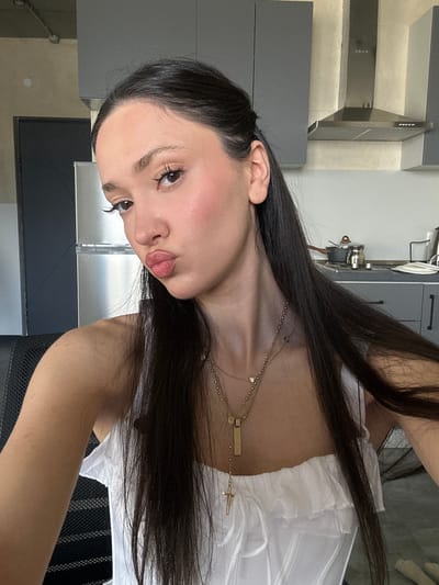 A selfie of a woman with long, dark hair wearing a white, ruffled top and a gold necklace with a cross pendant, set against a kitchen background. The kitchen features gray cabinets, a stainless steel refrigerator, and a cooker hood. The location is likely indoors. There is no information that allows me to confidently identify that the photo was taken in Zovuni, Armenia.
